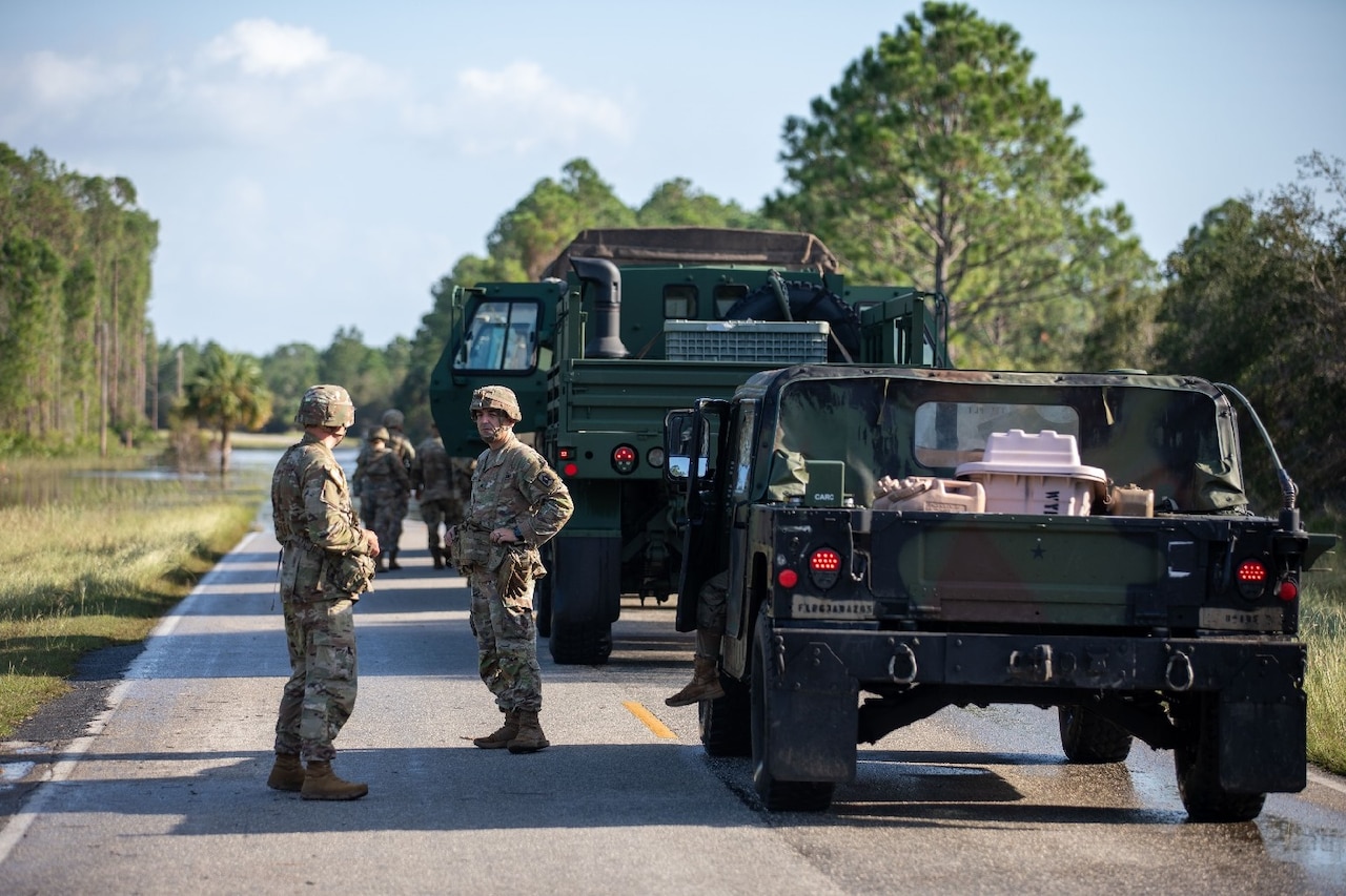 Service members in uniforms stand on a road next to military vehicles.