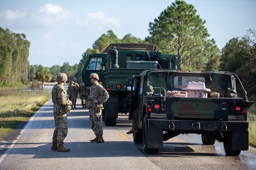 Service members in uniforms stand on a road next to military vehicles.