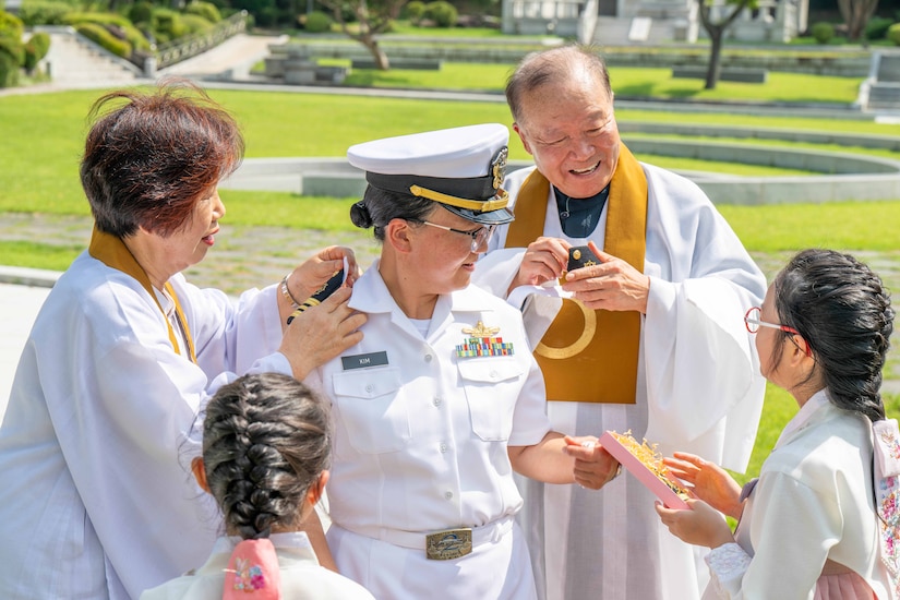 A Navy officer in a formal uniform is surrounded by family during a promotion ceremony on a sunny day.