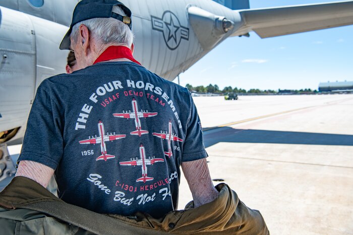 A man shows the back of his shirt that reads "THE FOUR HORSEMEN USAF DEMO TEAM, Gone But Not Forgotten" with four C-130 aircraft in formation in the center.