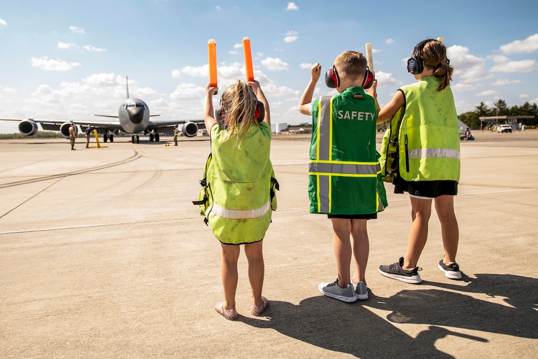 Three kids wearing neon vests holding batons signal an aircraft on a tarmac under a cloudy, blue sky.