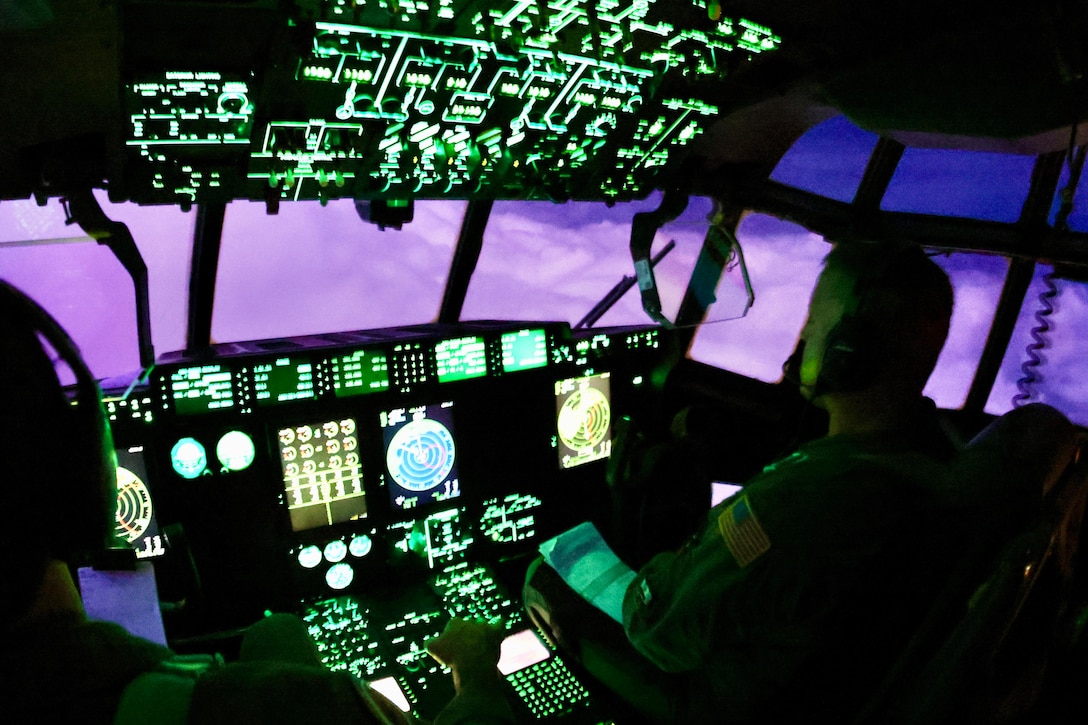 Pilots sit in an airborne cockpit illuminated by green gadgets as clouds are seen in a dark sky through the window.