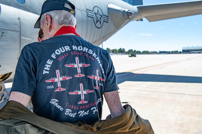 A man shows the back of his shirt that reads "THE FOUR HORSEMEN USAF DEMO TEAM, Gone But Not Forgotten" with four C-130 aircraft in formation in the center.