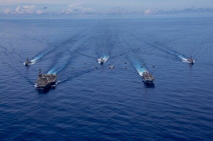 A view from the air of five large warships and several smaller vessels sailing in formation in the ocean.