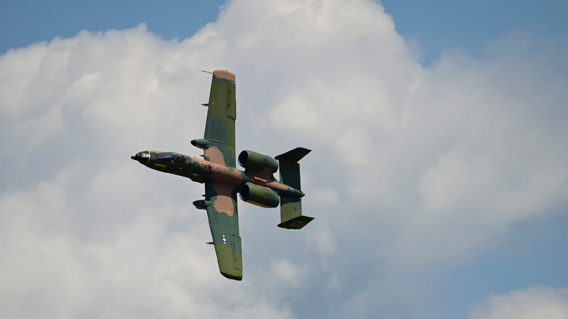 A military aircraft is flying in the sky with puffy clouds in the background.