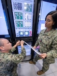 Two Navy personnel in uniform working at a computer server array, one on the phone.