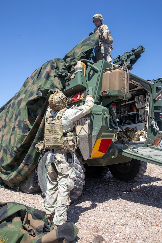 Two soldiers place a camo tarp over an armored military vehicle outside.