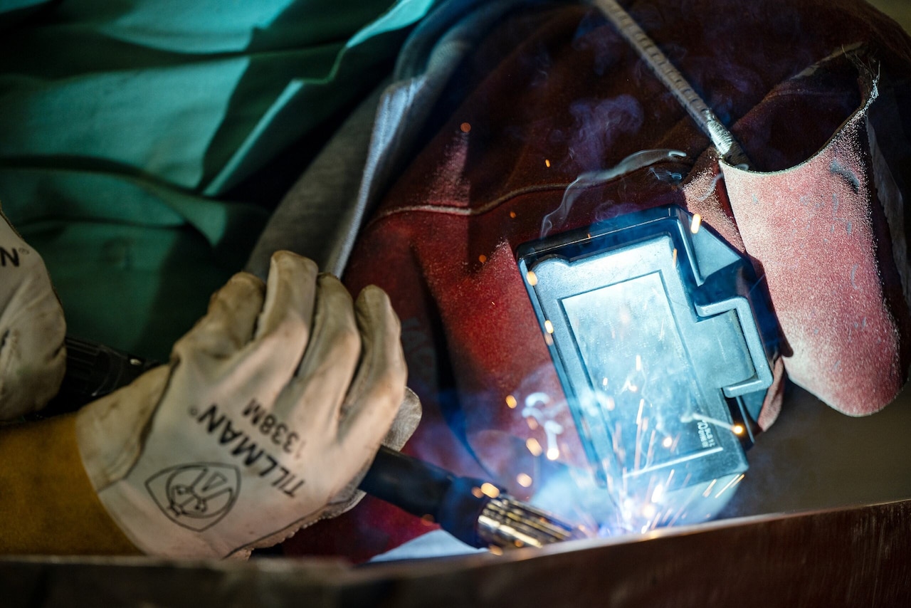 A person in a welding hood looks closely at a piece of steel as sparks shoot from the welding equipment.