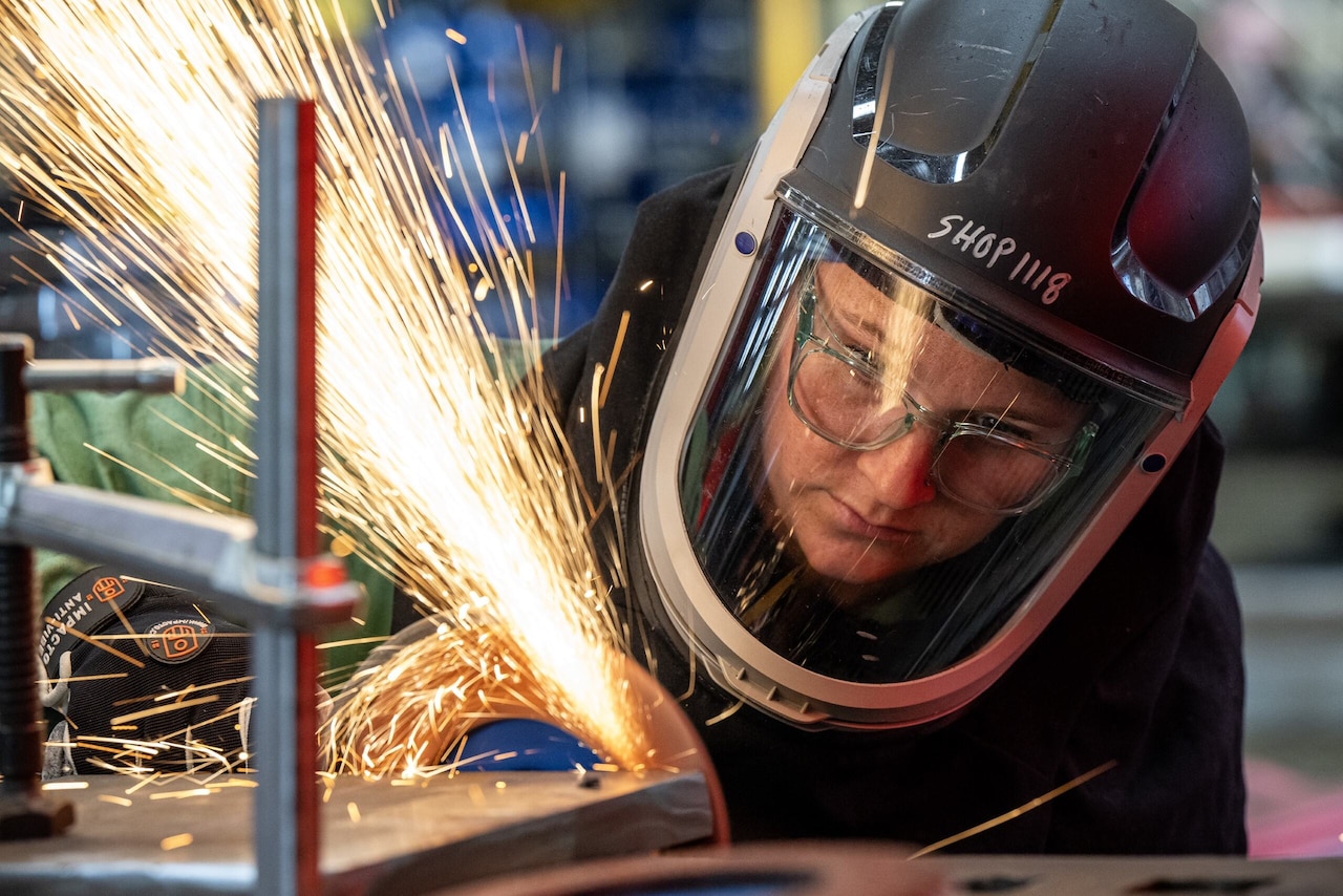 A person in a protective welding hood holds a piece of equipment against a piece of steel, causing sparks.