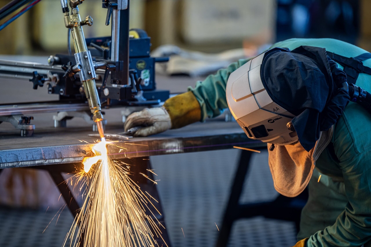 A person watches as an automated piece of equipment shoots sparks against a piece of steel.