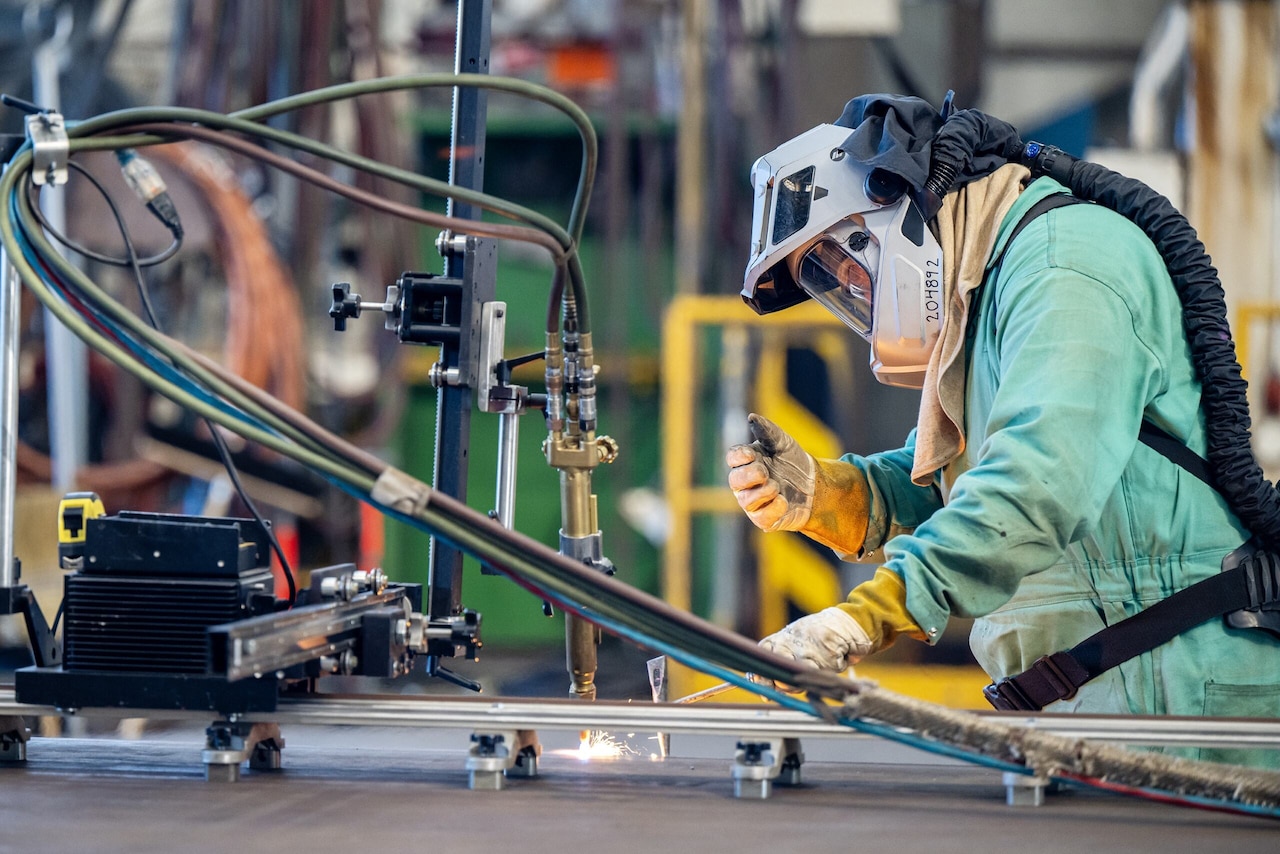 A person in protective gear adjusts a piece of welding equipment as sparks fly from it.