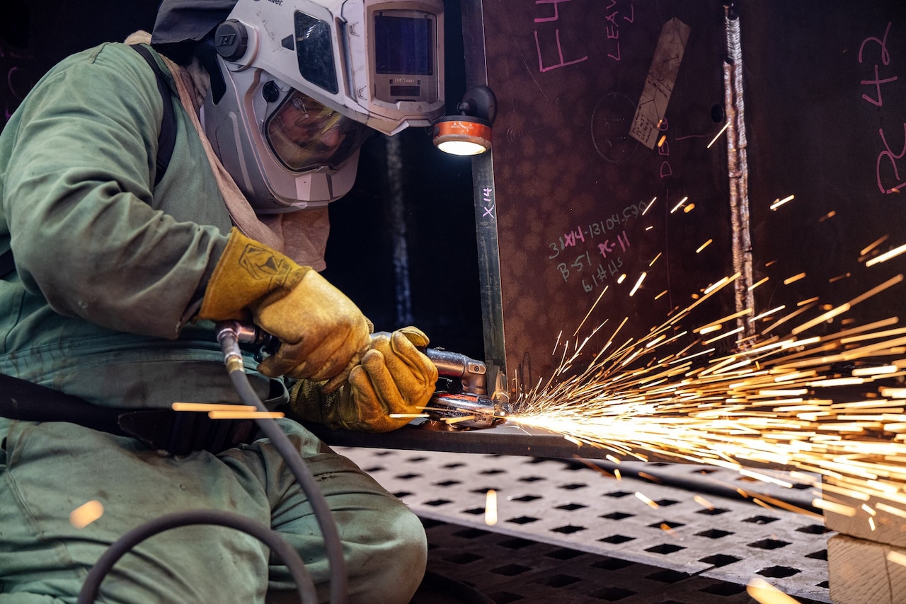 A person in a protective welding hood holds a piece of equipment against a piece of steel, causing sparks.