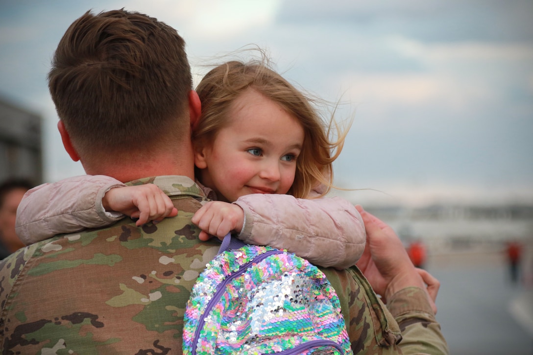A soldier hugs a smiling child who is holding a sparkly backpack.