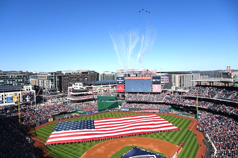 Guardsmen hold a giant flag on a baseball field with crowds in the stands and aircraft flying above.