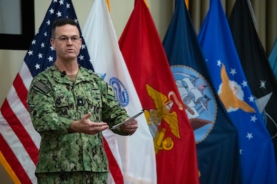 A man in a camouflage military uniform stands in front of American, U.S. Army, U.S. Marine Corps, U.S. Navy, U.S. Coast Guard and U.S. Space Force flags gesturing with his hands.