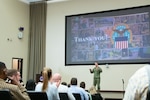 A man in a camouflage military uniform stands on stage in front of an audience in an auditorium. There is a display screen behind him that says 'thank you' with a photo collage and the Defense Logistics Agency logo.