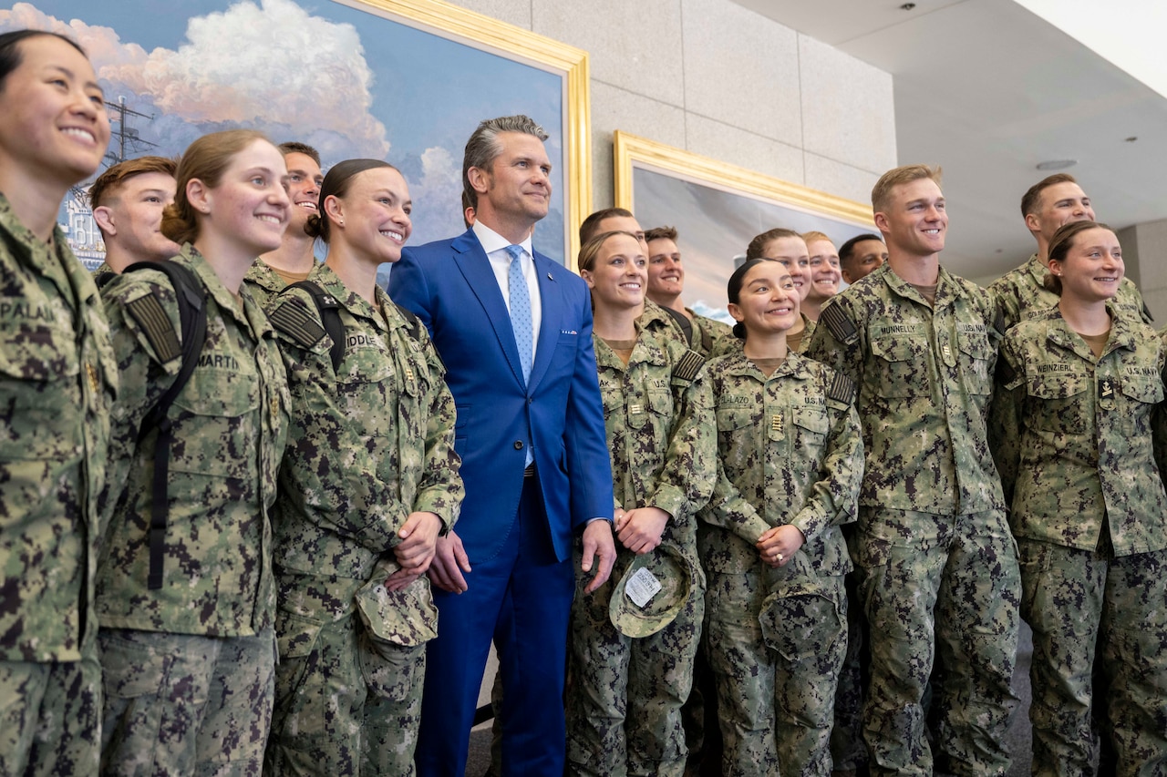 Defense Secretary Pete Hegseth is flanked by a group of midshipmen as they take a photo with paintings on the wall in the background.