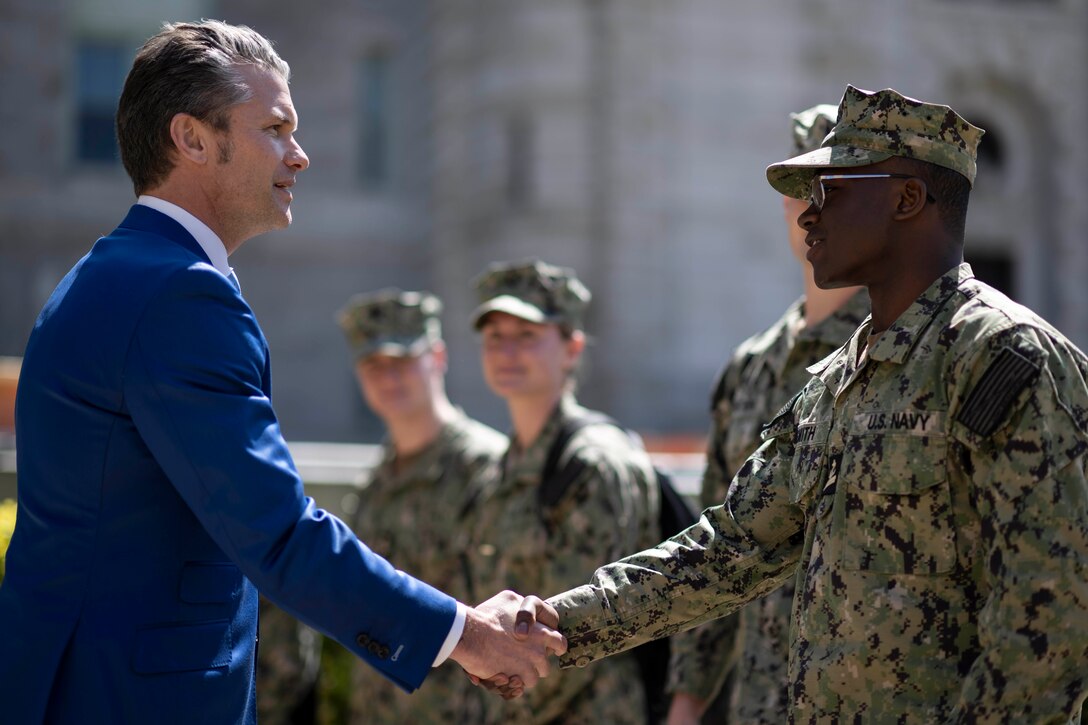 A man in a blue suit shakes hands with a midshipman in uniform while outdoors.