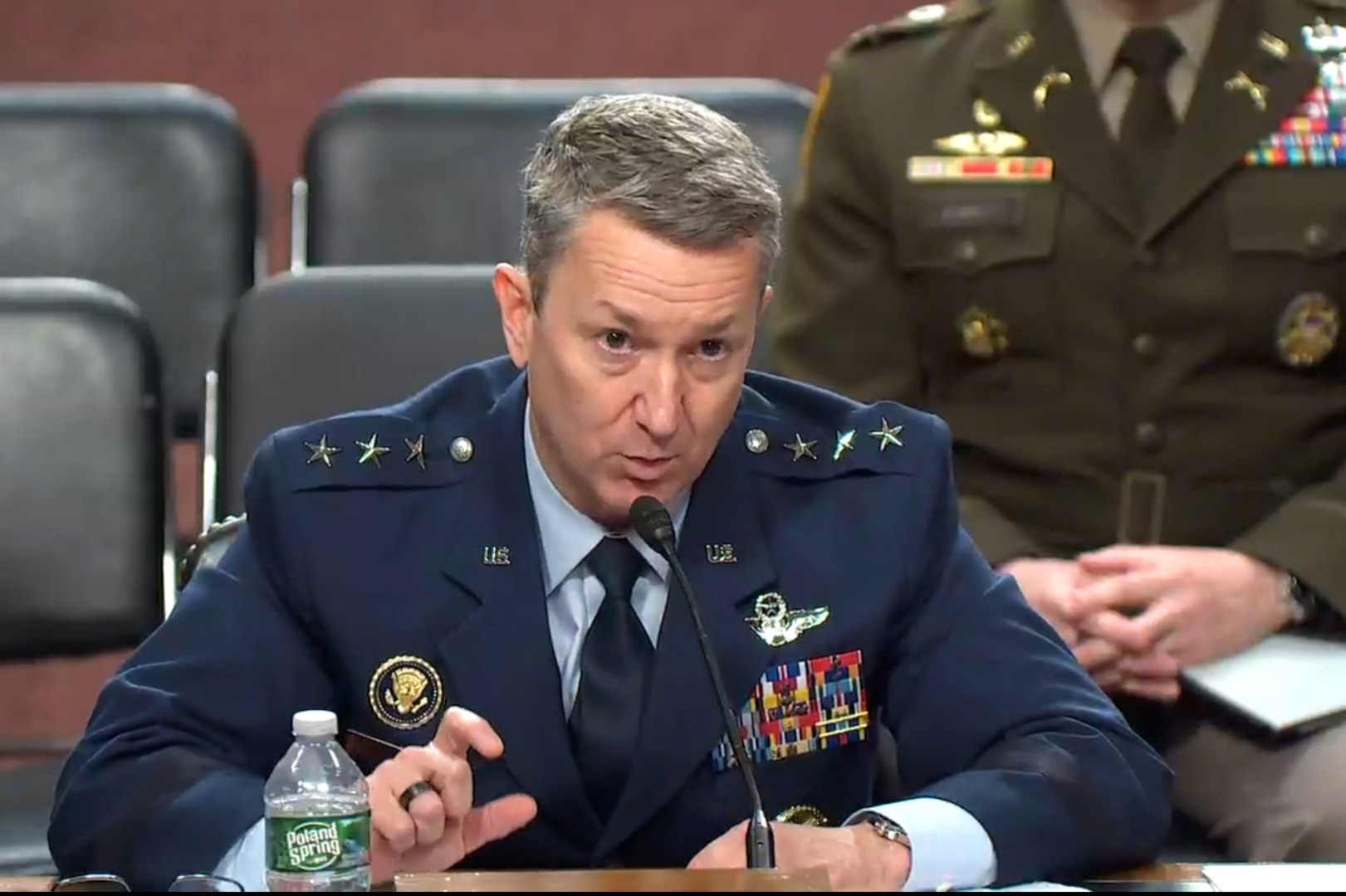 A man in a military uniform sits at a table behind a microphone.