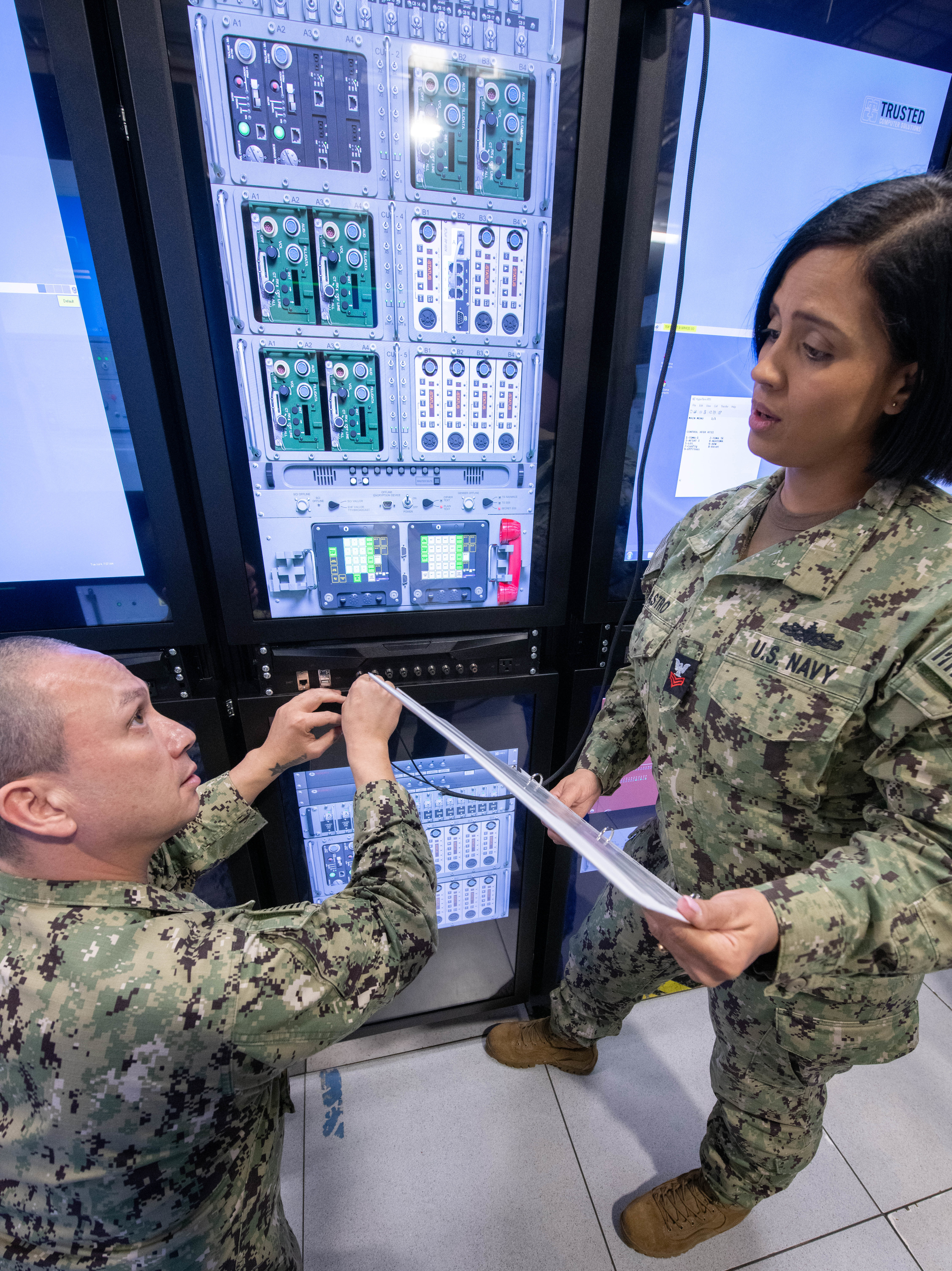 Two Navy personnel in uniform working at a computer server array, one on the phone.