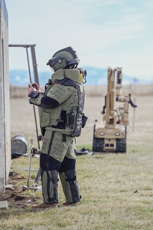 Man in EOD Bomb Suit scans building with phone with robot in the background