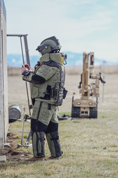 Man in EOD Bomb Suit scans building with phone with robot in the background