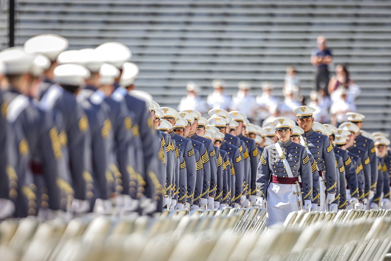Dozens of individuals in uniforms are lined up between folding chairs.