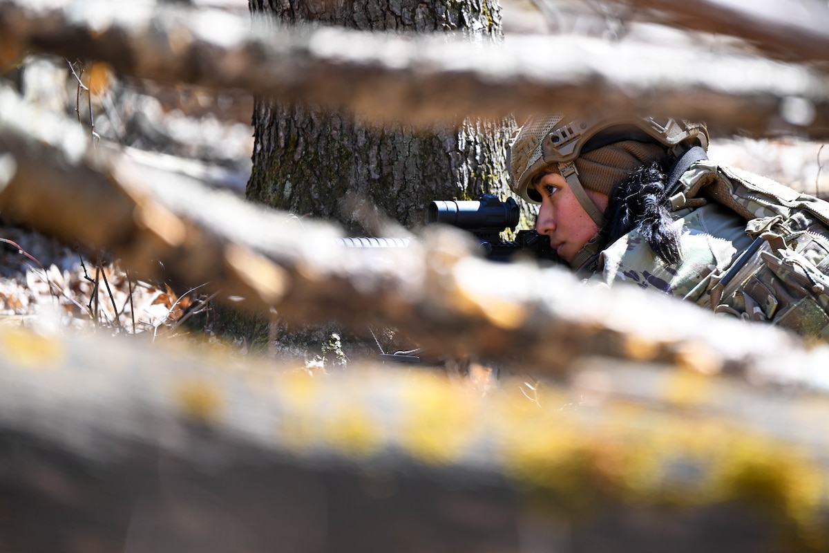 An airman looks through scope of a weapon while lying in a wooden area as seen through branches.