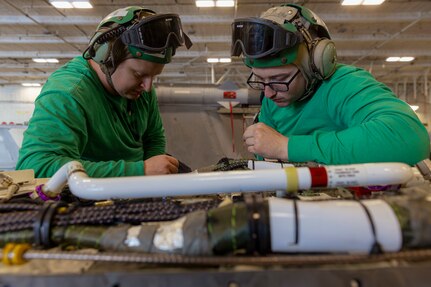 Aviation Electrician's Mate 1st Class John Clardy (left) and Aviation Electrician's Mate 2nd Class Phillip Newton, both  assigned to the "Gray Wolves" of Electronic Attack Squadron (VAQ) 142, re-pin the connector in an E/A-18G Growler in the hangar bay of the world's largest aircraft carrier, USS Gerald R. Ford (CVN 78), April 1, 2025.