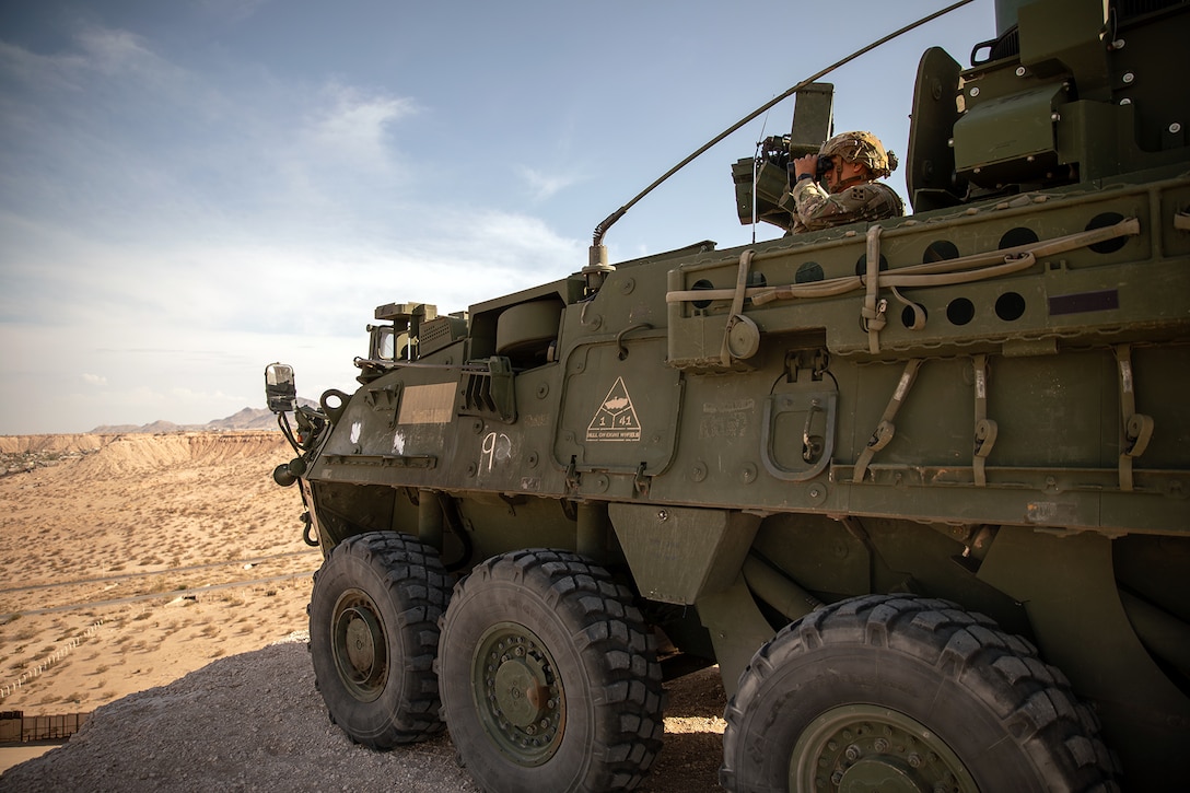 A soldier looks with binoculars from a military vehicle on sandy terrain.