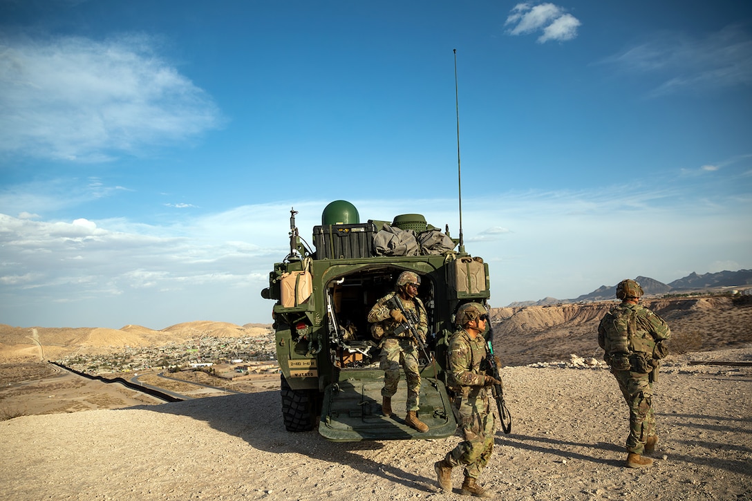 Soldiers exit a military vehicle onto sandy ground in an arid terrain.