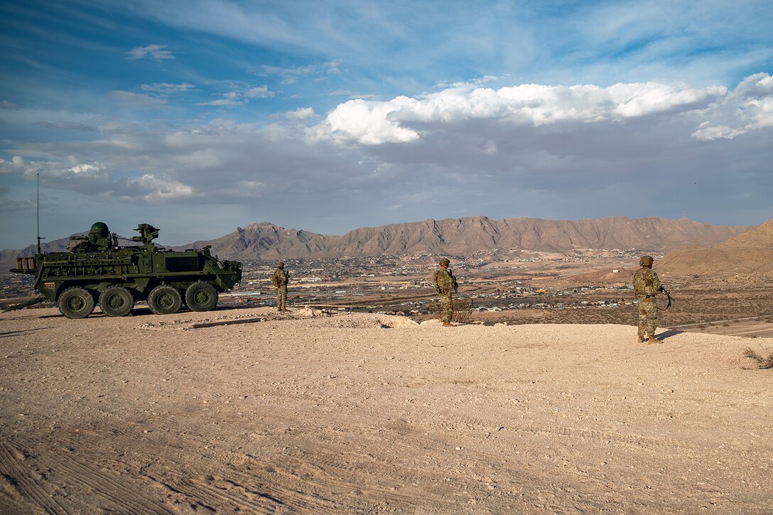 Soldiers stare out onto a plain next to a military vehicle.