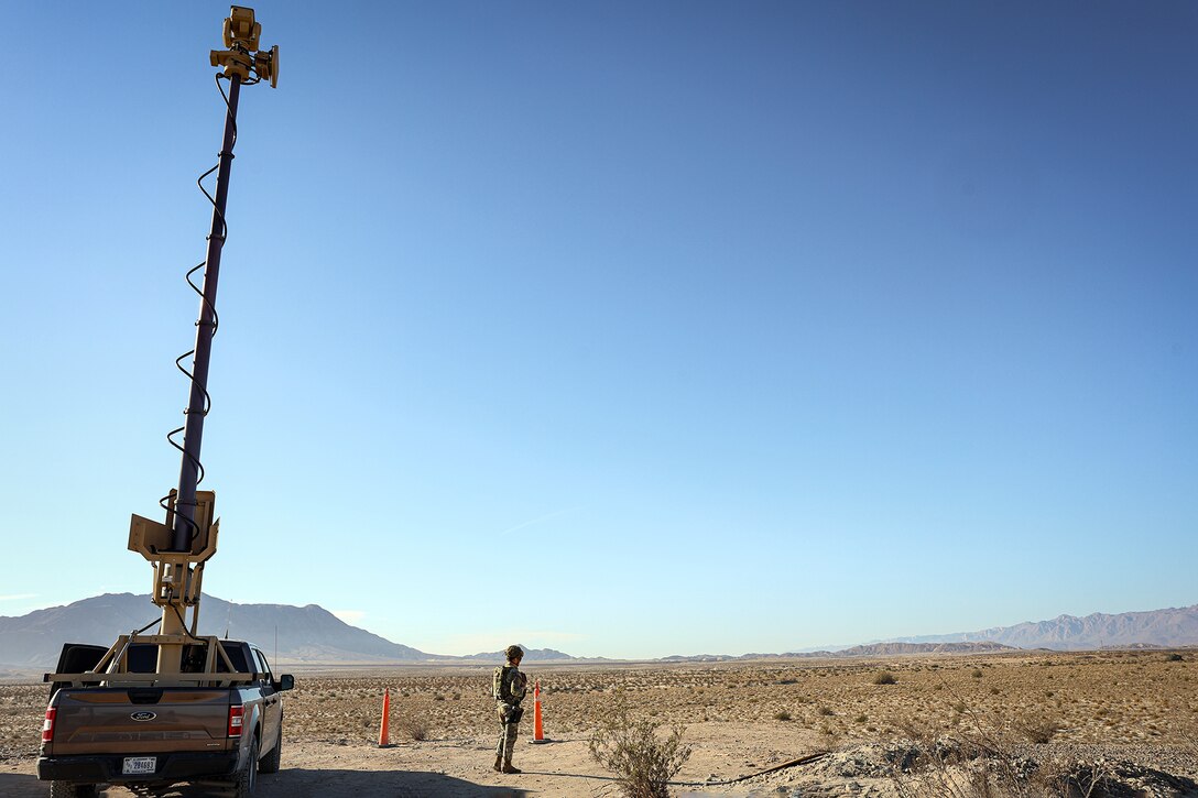 A soldier stands in an arid field next to a vehicle with a tall antenna.