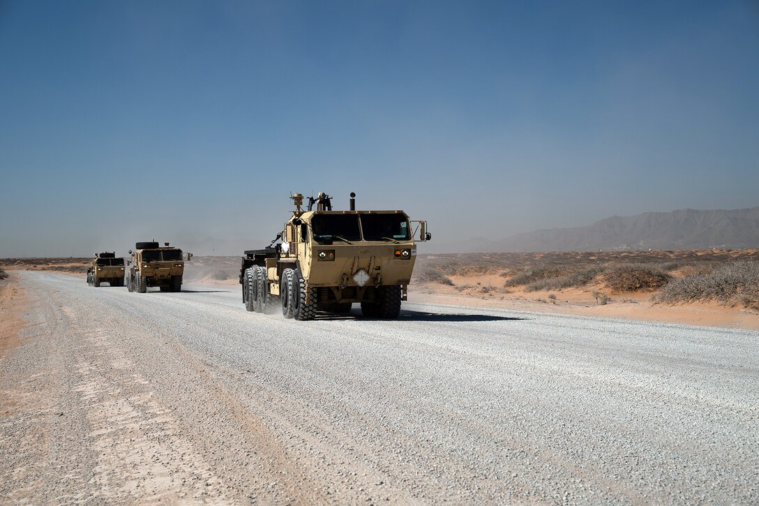 Three military vehicles drive down a dusty road on a clear day.