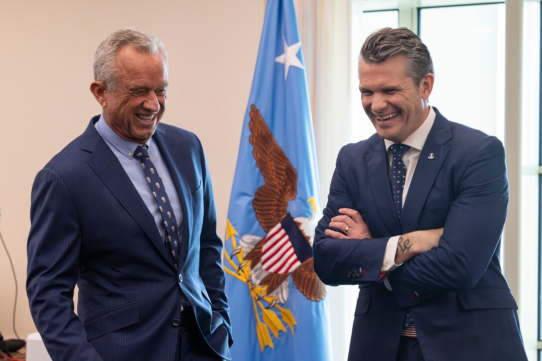 Two men wearing blue suits stand in front of a pentagon flag.