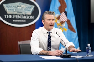 A man wearing a shirt and tie speaks into a microphone while sitting at a table in front of the Pentagon seal and flag.
