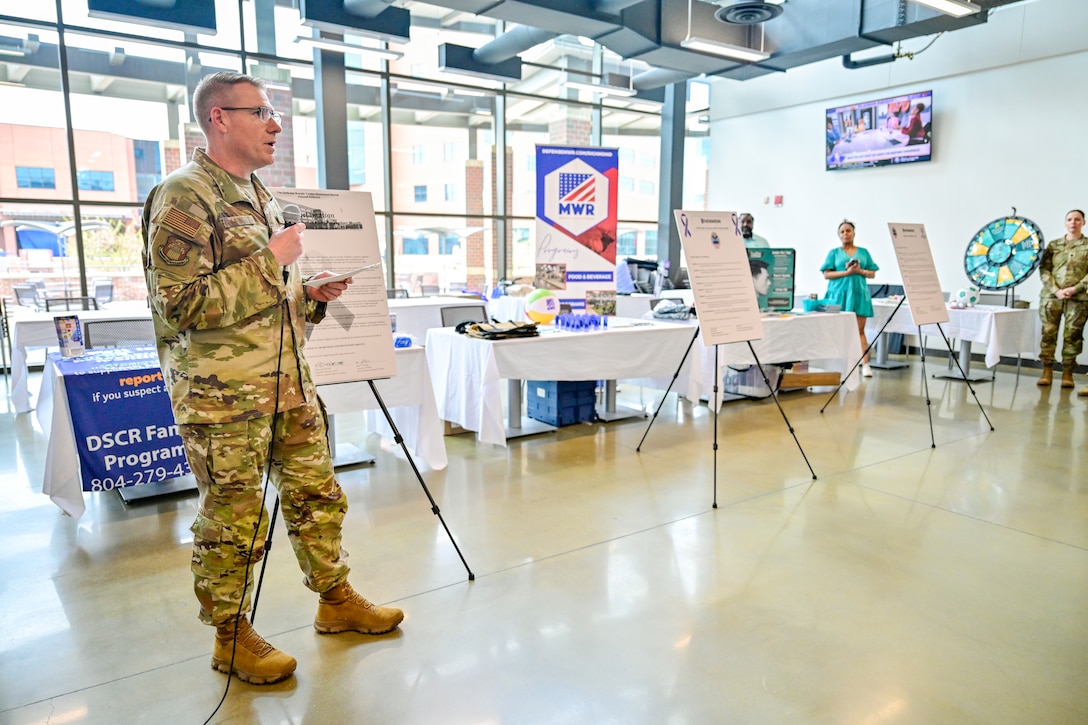A military officer holding a microphone stands next to a sign.