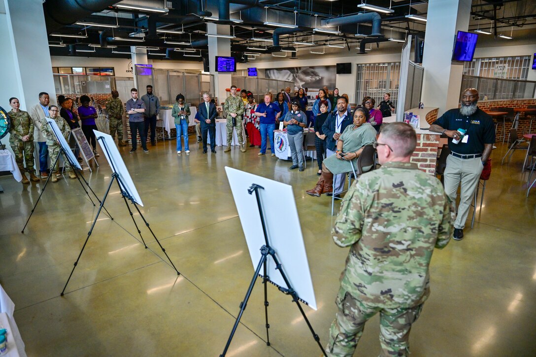 A group of employees listen to a military officer as he stands next to a sign.