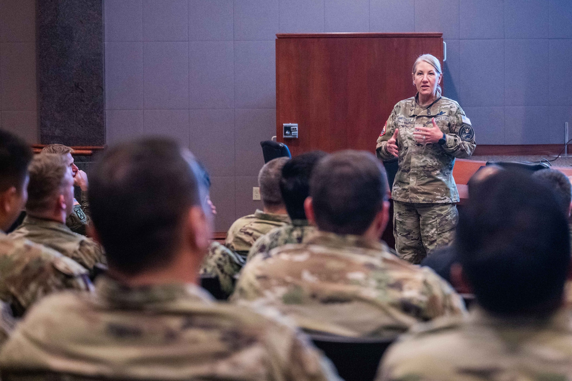 A military member speaks to a crowd of other military members in an auditorium.