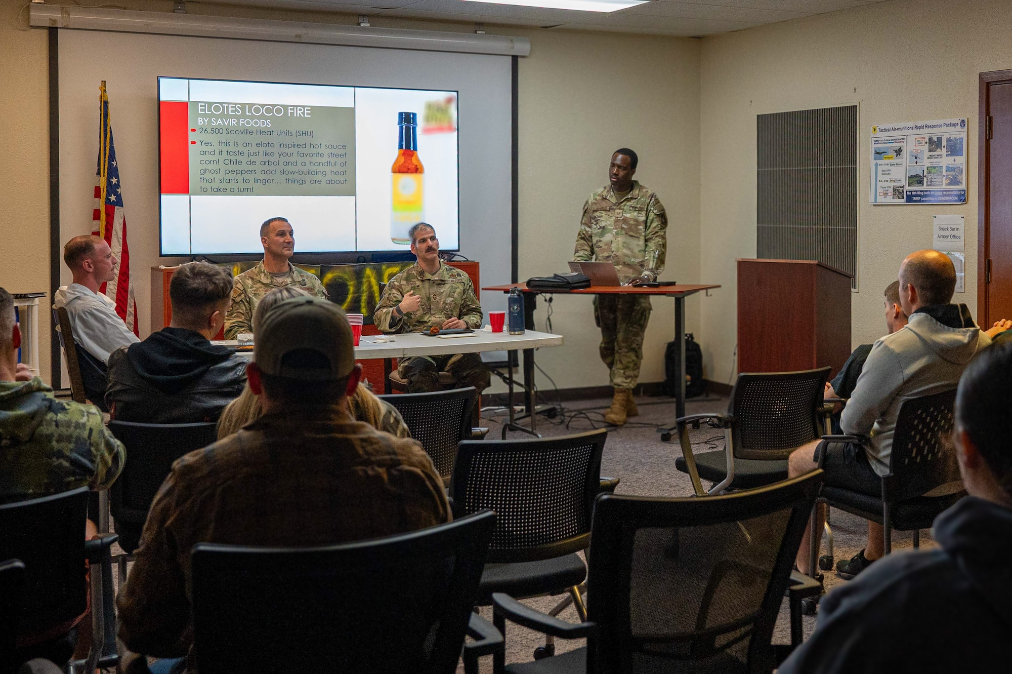 Three military members sit at a table eating chicken wings in front of a crowd
