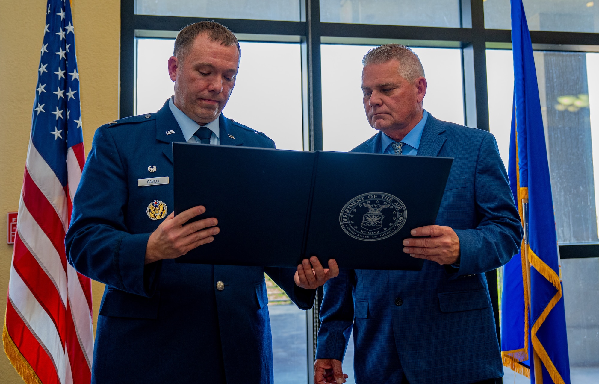 From left, a serviceman and a civilian man, standing in front of a large window with the sun coming through the panes, looks down at an award cover.