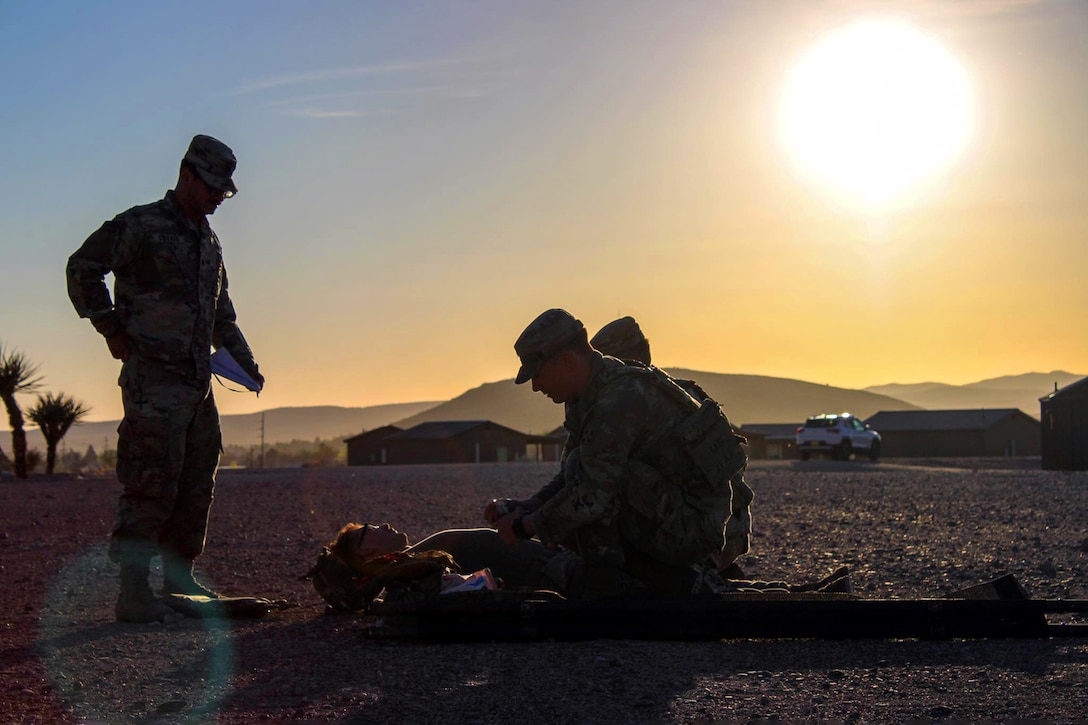 Soldiers stand and kneel next to someone laying down outside.