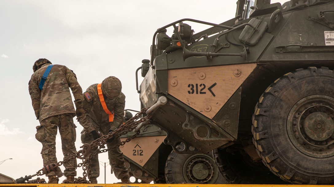 Two soldiers secure chains attached to a military vehicle.