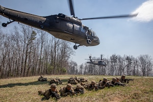 One row of soldiers lay in a field holding guns under two hovering helicopters.