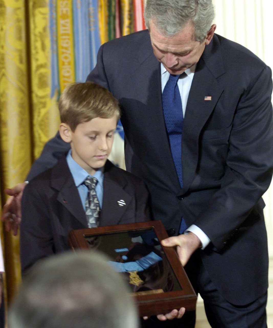 A man stands over a boy who is holding a shadow box that contains a medal.