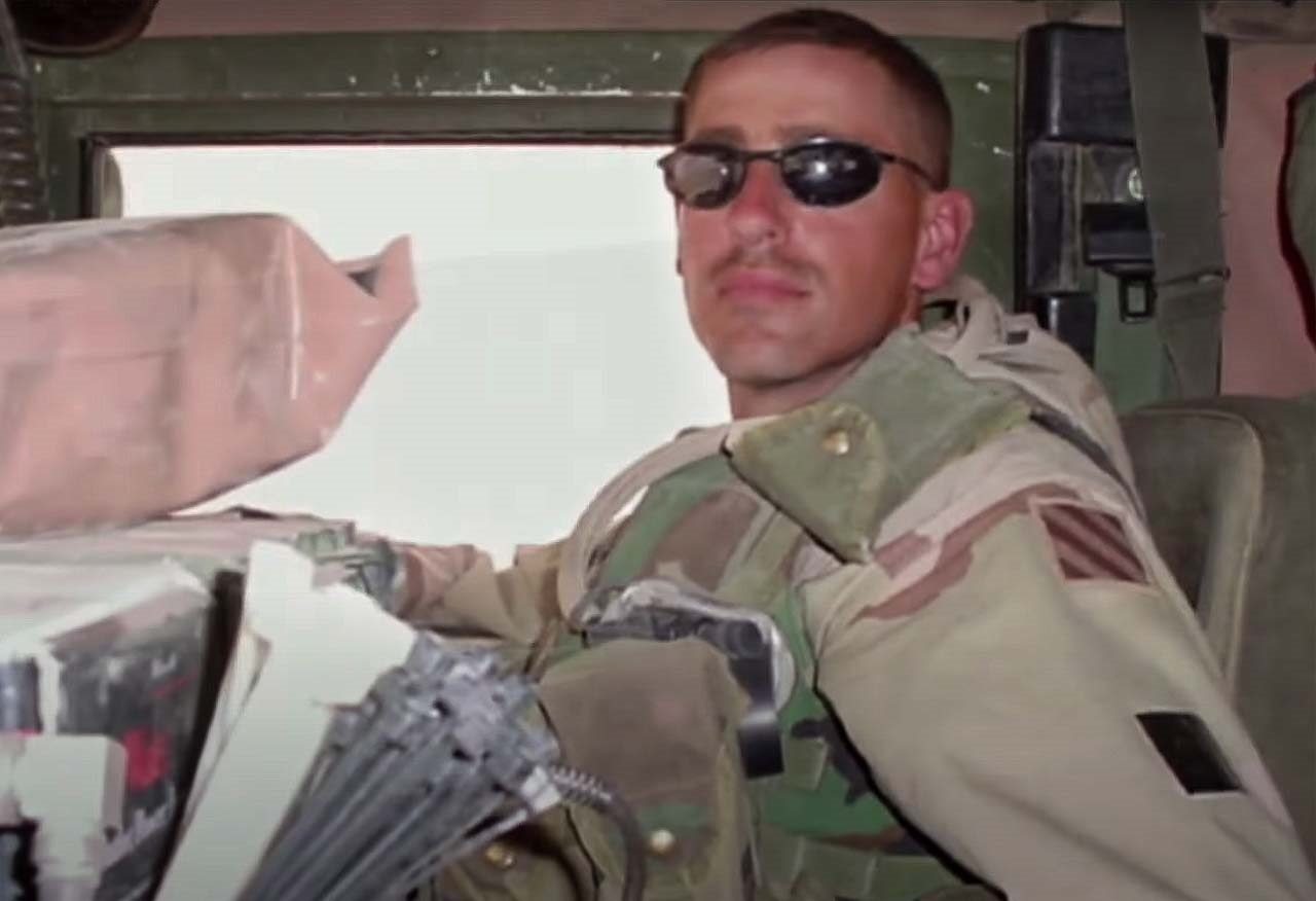 A man in combat fatigues and sunglasses sits in a vehicle.