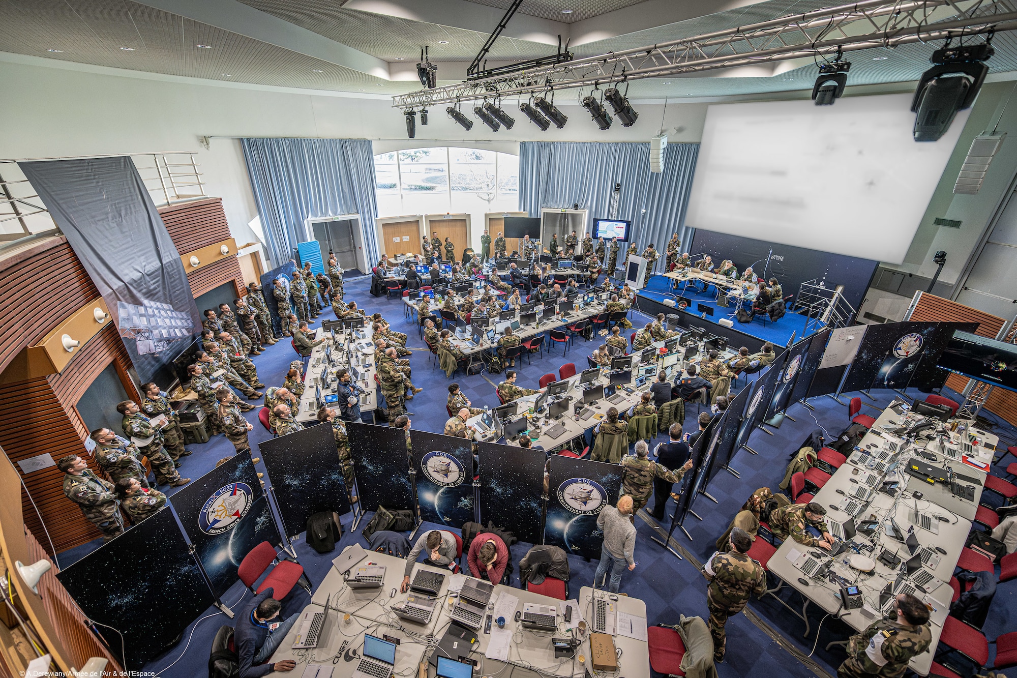 A wide photo of the operations floor during AsterX, France's premier space-focused military exercise Exercise participants are sitting and standing around the room.
