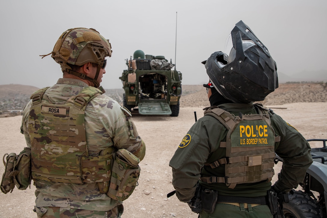 A soldier and a border patrol agent wearing a bike helmet chat on arid terrain as a military vehicle is parked in the background.