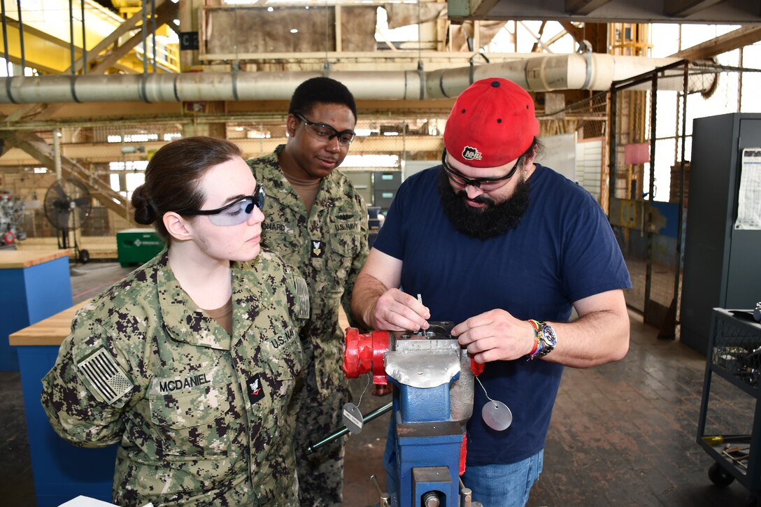 During their annual Navy Reserve two-week-long training period at Norfolk Naval Shipyard (NNSY), SurgeMain Charlotte Machinist Mate Third Class Ashton McDaniel (left) and SurgeMain Atlanta Machinist Mate First Class Kyle Leonard (center) watches NNSY’s Outside Machine Shop (Shop 38) Outside Machinist James Derrickson (right) as he demonstrates how to use a top-loaded ball valve, breaking down, taking measurements, using the center line tool to get the stem center line measurement and giving them a little refresher on some of the precision measuring tools that they would use and the proper tools to take apart the valve.