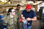During their annual Navy Reserve two-week-long training period at Norfolk Naval Shipyard (NNSY), SurgeMain Charlotte Machinist Mate Third Class Ashton McDaniel (left) and SurgeMain Atlanta Machinist Mate First Class Kyle Leonard (center) watches NNSY’s Outside Machine Shop (Shop 38) Outside Machinist James Derrickson (right) as he demonstrates how to use a top-loaded ball valve, breaking down, taking measurements, using the center line tool to get the stem center line measurement and giving them a little refresher on some of the precision measuring tools that they would use and the proper tools to take apart the valve.